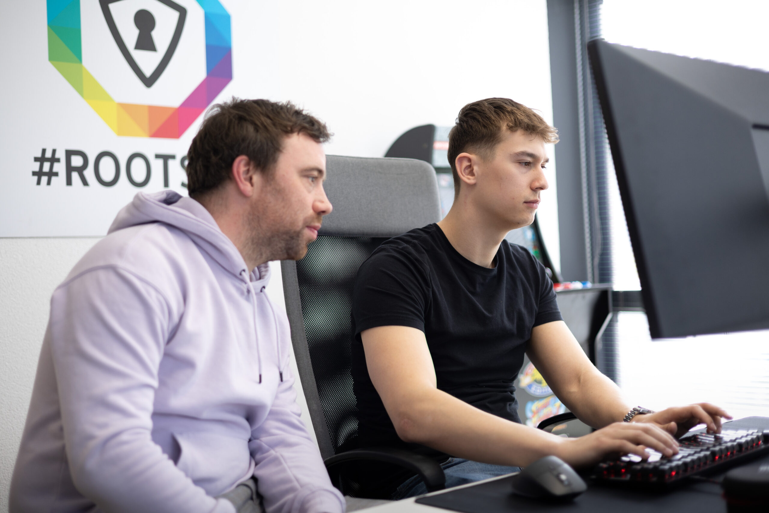 Two Rootsec colleagues working together at a workstation beneath the #ROOTSEC wall logo